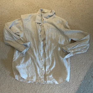 Beige linneskjorta - En stilren ljusbeige skjorta med klassisk krage och vita knappar hela vägen framtill. Skjortan har lång ärm och är tillverkad i ett lätt material som passar perfekt för en avslappnad look. 