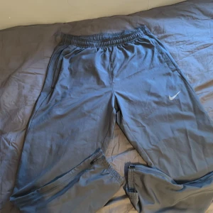 Grå träningsbyxor från Nike Dri-Fit - Säljer ett par grå Nike Dri-Fit träningsbyxor med resår i midjan och dragkedja vid fickorna. Byxorna har en klassisk Nike-logga på benet och är tillverkade i ett lätt syntetmaterial som andas.