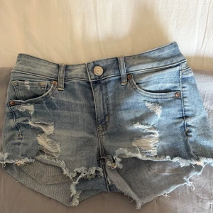Ljusblå slitna jeansshorts - Ljusblå lågmidjade jeansshorts med råa, fransiga kanter och slitningar framtill. Klassisk femficksmodell med knapp och dragkedja. Tillverkade i bomull med lite stretch för extra komfort. Perfekta för sommardagar och en avslappnad stil. Har sytt in dom i midjan