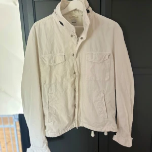 Vit jacka från Aspesi med dragkedja - Säljer en stilren vit jacka från Aspesi med dragkedja och knappar framtill. Jackan har två bröstfickor med lock och två snedställda fickor nertill. Hög krage och justerbar nederkant med snörning. Perfekt för sommargarderoben. Storleken är L, men de italienska jackorna sitter mindre. Skulle själv säga S eller M storleksmässigt. Nypris ca 6000kr