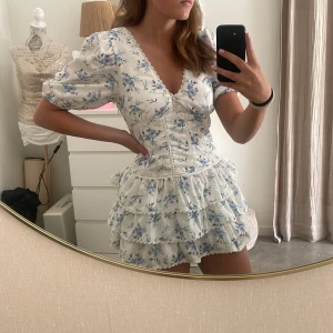 Vit blommig kortklänning med volanger - Superfin vit kortklänning med blått blommönster och volanger. Klänningen har puffärmar, v-ringning med spetsdetaljer och markerad midja. Perfekt för sommarens alla tillfällen! Inga defekter men har en liten fläck vid kragen som försvinner i tvätten☺️hör av er vid frågor 