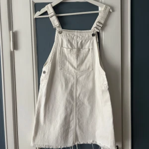 Vit jeansklänning med hängslen - Säljer en vit jeansklänning med klassiska hängslen och stor ficka framtill. Klänningen har justerbara axelband och rå kant nertill för en avslappnad look. Perfekt att styla med en t-shirt eller tröja under.