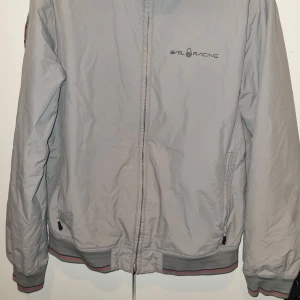 Ljusgrå vindjacka från Sail Racing - Ljusgrå vindjacka med dragkedja från Sail Racing. Jackan har ribbade muddar med röd och mörkgrå detalj, två fickor med dragkedja och broderad logga på bröstet. Perfekt för blåsiga dagar.