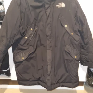 Svart vinterjacka från The North Face - Säljer en svart vinterjacka från The North Face med dragkedja och knappar. Jackan har flera fickor framtill och klassisk logga på bröstet. Perfekt för kalla dagar och har lång ärm för extra värme.