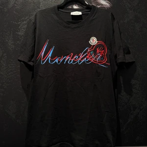 Svart t-shirt från Moncler med broderad logga - Svart t-shirt från Moncler med stort broderat Moncler-märke och färgglad logotyp framtill. Klassisk passform och rund halsringning. Perfekt för en stilren och trendig look.