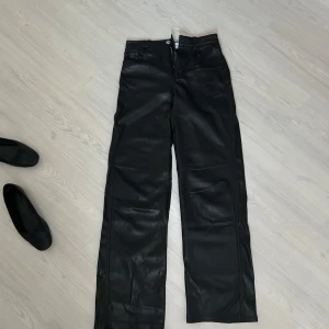 Svarta skinnbyxor från Bershka - Säljer ett par svarta skinnbyxor från Bershka med normal passform och raka ben. Byxorna har hög midja, klassisk femficksdesign och stängs med dragkedja och knapp. 