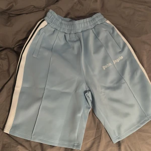 Ljusblå shorts från Palm Angels - Snygga ljusblå shorts från Palm Angels med vita sidoränder och broderad logga på vänster ben. De har elastisk midja med snörning och två fickor framtill. Perfekta för en avslappnad och sportig stil.