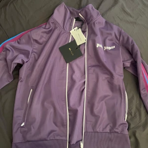 Lila träningsbyxor från Palm Angels - Säljer ett helt ny oanvänd tracksuit från Palm Angels med vita loggan på framsidan. Byxorna har elastisk midja och ränder i blått och rosa längs sidorna. Samt koftan Perfekta för en sportig och trendig look. Äkta. Kan sälja enskild det bara och fråga :)