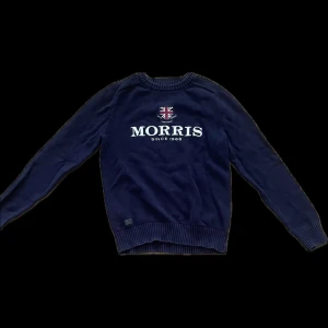 Mörkblå tröja från Morris Gents - Mörkblå långärmad tröja från Morris Gents med rund halsringning. Framsidan har ett tryckt Morris-logo och brittisk flagga. Tröjan är ribbad vid ärmslut och nederkant, perfekt för en stilren och klassisk look.
