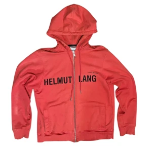 helmut lang hoodie by shayne oliver - Säljer en röd hoodie från Helmut Lang med svart logotyptryck över bröstet. Tröjan har dragkedja framtill, huva med dragsko och två fickor. Tillverkad i 100% bomull för en skön och avslappnad känsla. Made by 