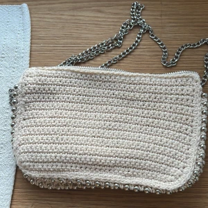 Beige stickad axelväska från Zara - Säljer en beige stickad axelväska från Zara med silverfärgad kedja och dragkedja. Väskan har virkade detaljer och metallpärlor längs kanterna. Perfekt för att bära dina viktigaste saker med stil.