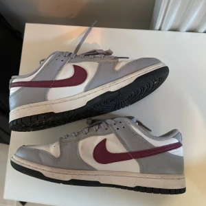 Nike Dunk Low med grå och vinröda detaljer - Säljer ett par Nike Dunk Low sneakers i vitt och ljusgrått med vinröd swoosh. Skorna har klassisk låg modell, snörning och platt sula. Perfekta för dig som gillar streetwear och vill ha en stilren men ändå färgstark sneaker.
