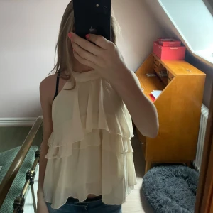 Beige volangtopp - Säljer en luftig beige topp med flera volanglager och lös passform. Toppen är ärmlös, perfekt för varma dagar eller när du vill ha något lätt och snyggt. Aldirig använd!🥰