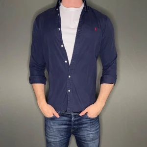 Mörkblå skjorta från Ralph Lauren - Snygg mörkblå skjorta från Ralph Lauren med klassisk krage och knappar framtill. Liten röd broderad logga på bröstet. Skjortan har lång ärm och normal passform, perfekt att bära öppen över en t-shirt.