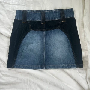 Jeanskjol från Alwear med breda hällor - Säljer en cool jeanskjol från Alwear i klassisk blå denim. Kjolen har breda bälteshällor, nitar framtill och kontrasterande mörka partier på sidorna. Perfekt för dig som gillar en edgy och trendig stil.