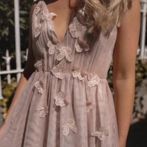 Ljusrosa långklänning med blommiga detaljer - Romantisk ljusrosa långklänning med v-ringning och blommiga spetsapplikationer. Klänningen är ärmlös och har ett luftigt, svepande tyg som ger en elegant känsla. Perfekt för speciella tillfällen. Är i en M och har en jätte liten fläck under en av blommorna men som knappt syns. Säljer då den inte passade mig❤️