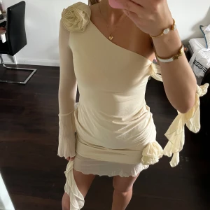 Creamvit  one shoulder-klänning med volangdetaljer - Superfin kortklänning i champagne/cream med one shoulder-design och stora blomdetaljer på axel och höft. Klänningen har en figurnära passform och dekorativa rosetter samt volangkant nertill.