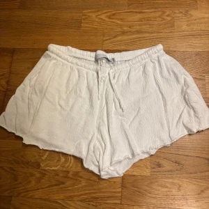 Vita shorts med dragsko - Säljer ett par vita shorts i mjukt och lätt material med resår och dragsko i midjan. De har en lös passform och vågiga kanter nertill, perfekta för varma dagar.