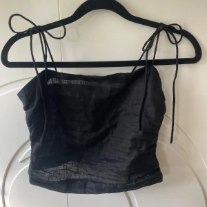 Svart linnetopp med knytband från H&M - Snygg svart linnetopp från H&M med tunna axelband som knyts på axlarna. Toppen är croppad och har smockad rygg för extra komfort. Perfekt till varma dagar och enkel att matcha med olika outfits.