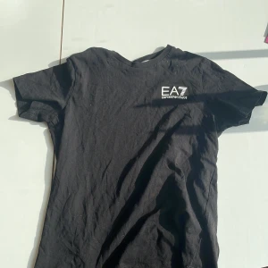 Svart t-shirt från Emporio Armani EA7 - Säljer en svart t-shirt från Emporio Armani EA7 med vit logga tryckt på bröstet. Klassisk rund hals och korta ärmar. Perfekt för en stilren och sportig look.  Jag säljer T-shirten för att den växt ut mig pris kan dyskutwras