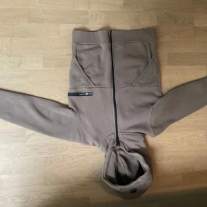 Beige fleecejacka med huva från Sail Racing - Mysig beige fleecejacka från Sail Racing med huva och hel dragkedja framtill. Jackan har två fickor och en svart logodetalj på bröstet. Perfekt för kyliga dagar och enkel att matcha med det mesta.