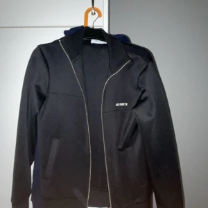 Svart jacka med dragkedja från Les Deux - Säljer en svart tröja från Les Deux med stilren design och silverfärgad dragkedja. Jackan har lång ärm och en enkel, sportig look med diskret logga på bröstet. Perfekt för dig som gillar minimalistisk stil. Perfekt på sommarkvällar och sådant , helt oanvänd stil s