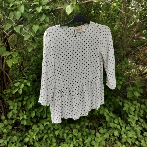 Vit blus med svarta prickar - Säljer en söt vit blus med svarta prickar och volang nertill. Blusen har rund halsringning och trekvartsärm. Perfekt för dig som gillar lekfulla mönster och en luftig känsla.