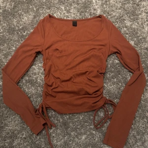 Rostbrun ribbad topp med knyt - Säljer en rostbrun långärmad topp med ribbad struktur och knyt i sidorna. Toppen har en rundad urringning och är figurnära i modellen. Perfekt för dig som gillar trendiga detaljer och snygg passform. Storlek S men sitter tajt så mer åt Xs/S. Använd 1 gång och har tyvärr ingen bra bild. 