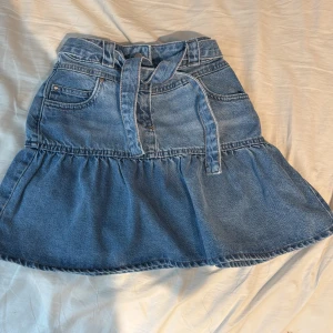 Jeanskjol med volang och bälte från Next - Säljer en blå jeanskjol från Next med volang nertill och tillhörande bälte i midjan. Kjolen har fickor fram och bak samt justerbar midja för perfekt passform. Perfekt för dig som gillar denim och vill ha en söt och trendig look. På bild är den justerad till minsta storlek och är menat för yngre ungdom. Den går att göra större och är aldrig använt med etikett kvar, köparen står för frakt 💗