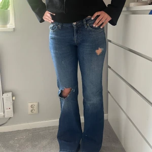 Low waist bootcut med slitningar - Blå lågmidjade jeans med hål på benen och bakficka. Används ca 2 gånger. Storlek 26/32