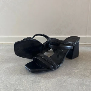 Svarta sandaler med klack och glittriga band - Säljer ett par svarta sandaler med bred klack och två smala glittriga remmar över foten. Skorna har fyrkantig tå och öppen häl, vilket ger en trendig look. Perfekta för att lyfta en outfit med lite extra bling.