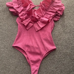 Rosa volangbody - Säljer en rosa body med stora volangdetaljer runt v-ringningen. Bodyn är ärmlös och har en ribbad struktur i tyget. Perfekt för dig som vill sticka ut med en färgglad och trendig topp.