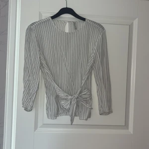 Vit och svart randig blus från Stradivarius - Säljer en vit blus med svarta ränder från Stradivarius. Blusen har nästan hel långa långa ärmar, knytning i midjan och en liten knapp i nacken. 