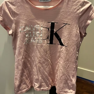 Rosa t-shirt från Calvin Klein Jeans - Säljer en ljusrosa t-shirt från Calvin Klein Jeans med stor svart CK-logga och silvrig text på bröstet. T-shirten har rund halsringning och korta ärmar. Perfekt för en avslappnad och trendig look.