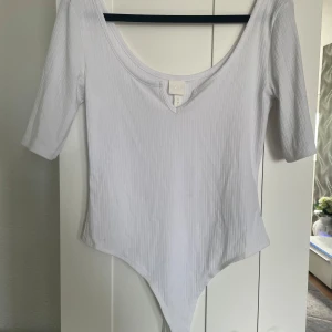 Vit ribbad body från H&M - Säljer en vit ribbad body från H&M med djup rundad ringning och trekvartsärm. Perfekt att styla med jeans eller kjol för en enkel och stilren look.