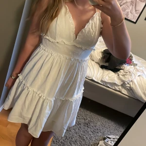 Vit kortklänning - Supersöt vit klänning med smala axelband och v-ringning. Klänningen har volangdetaljer och ett diskret prickigt strukturmönster i tyget. Perfekt till skolavslutningar och till sommaren 