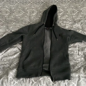 Mörkgrå hoodie från Ralph Lauren - Mörkgrå hoodie med dragkedja från Ralph Lauren. Tröjan har broderad logga på bröstet, två fickor framtill och justerbar huva med snören. Perfekt för en avslappnad stil. Lite knottrig därför säljer jag den lite billigare. Hör av er vid frågor