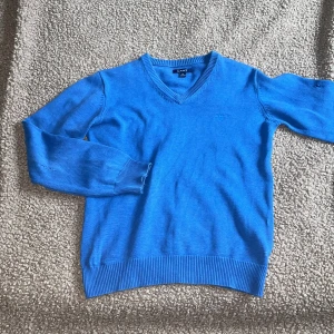 Blå v-ringad tröja från GANT - Säljer en klassisk blå v-ringad tröja från GANT med broderad logga på bröstet. Tröjan har långa ärmar och ribbade muddar vid ärmslut och nederkant. Perfekt för dig som gillar stilrena och tidlösa plagg.