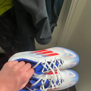  fotbollsskor från Adidas - Säljer ett par snygga fotbollsskor från Adidas i vitt, blått och rött. Skorna har snörning och en sportig design med röda detaljer på sidorna och blå insida. Perfekta för dig som vill sticka ut på planen! De är aldrig använda så de är i riktigt bra pris
