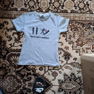 Grå t-shirt med tryck 'How to drop a boyfriend' - Grå t-shirt med svart och rött tryck på bröstet som visar en humoristisk illustration och texten 'How to drop a boyfriend'. Klassisk rund hals och korta ärmar. Perfekt för dig som gillar roliga statement-plagg.