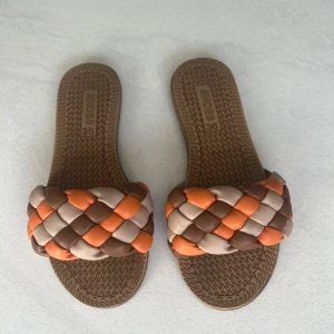 Flätade sandaler i brunt, beige och orange - Säljer ett par sandaler med flätad rem i brunt, beige och orange. Skorna har platt sula och öppen tå, perfekta för varma dagar. Snygg och trendig design som sticker ut!