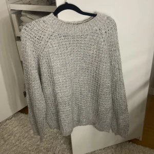Ljusgrå stickad tröja - Mysig ljusgrå stickad tröja med grovstickat mönster och rund halsringning. Perfekt för kyliga dagar och enkel att matcha med olika outfits. Långa ärmar och avslappnad passform.