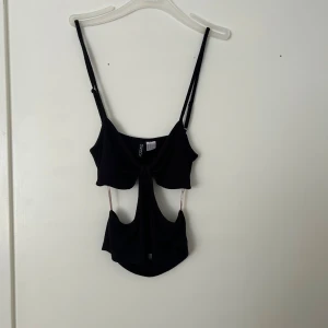 Svart croppad cut-out topp - Trendig svart croppad topp med cut-out detaljer och smala axelband. Toppen har en unik design med öppningar framtill och passar perfekt till en snygg outfit.