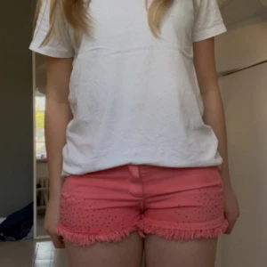 Rosa/korall shorts  - Säljer ett par korallrosa shorts med fransig kant och små silvriga nitar framtill. Shortsen har normal passform, knapp och dragkedja samt fickor både fram och bak. Perfekta för sommaren! Finns en lite fläck på baksidan men syns knappt, se sista bilden. 39 cm i midjemått 