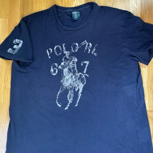 Polo Ralph Lauren T-Shirt - Säljer en riktigt fet mörkblå t-shirt från Polo Ralph Lauren med stort tryck på framsidan. Siffran 3 är tryckt på ena ärmen. Klassisk rund hals och L passform. 100% bomull. Perfekt nu inför sommaren med snygga linnebyxor eller shorts!
