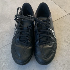 Svarta Nike Tiempo fotbollsskor - Säljer ett par svarta Nike Tiempo fotbollsskor med vit swoosh och detaljer. Skorna har snörning och är tillverkade i syntetmaterial. Perfekta för dig som vill ha stilrena och klassiska fotbollsskor på planen.