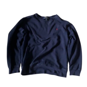 Mörkblå sweatshirt från Polo Ralph Lauren - Mörkblå sweatshirt från Polo Ralph Lauren med klassisk rund halsringning och broderad röd logga på bröstet. Tröjan har långa ärmar och är tillverkad i mjuk bomull, perfekt för en avslappnad stil.