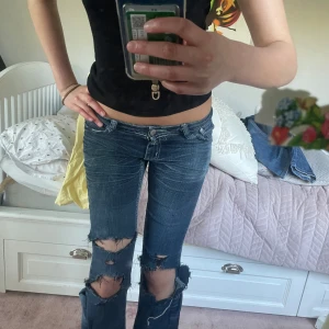 Blå slitna bootcut jeans - Säljer ett par blå bootcut jeans med låg midja och slitna detaljer på knäna. .