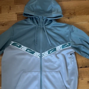 Ljusblå hoodie från Nike - Säljer en snygg ljusblå hoodie från Nike med dragkedja och huva. Jackan har en vit och grön rand med Nike-logga över bröstet och två fickor framtill. Perfekt för en sportig stil.
