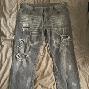 Grå slitna jeans från DSQUARED2 - Grå jeans från DSQUARED2 med slim fit och tydliga slitningar samt färgstänk på både fram- och baksida. Byxorna har klassisk femficksdesign och dragkedja. Märket syns tydligt på insidan av linningen.